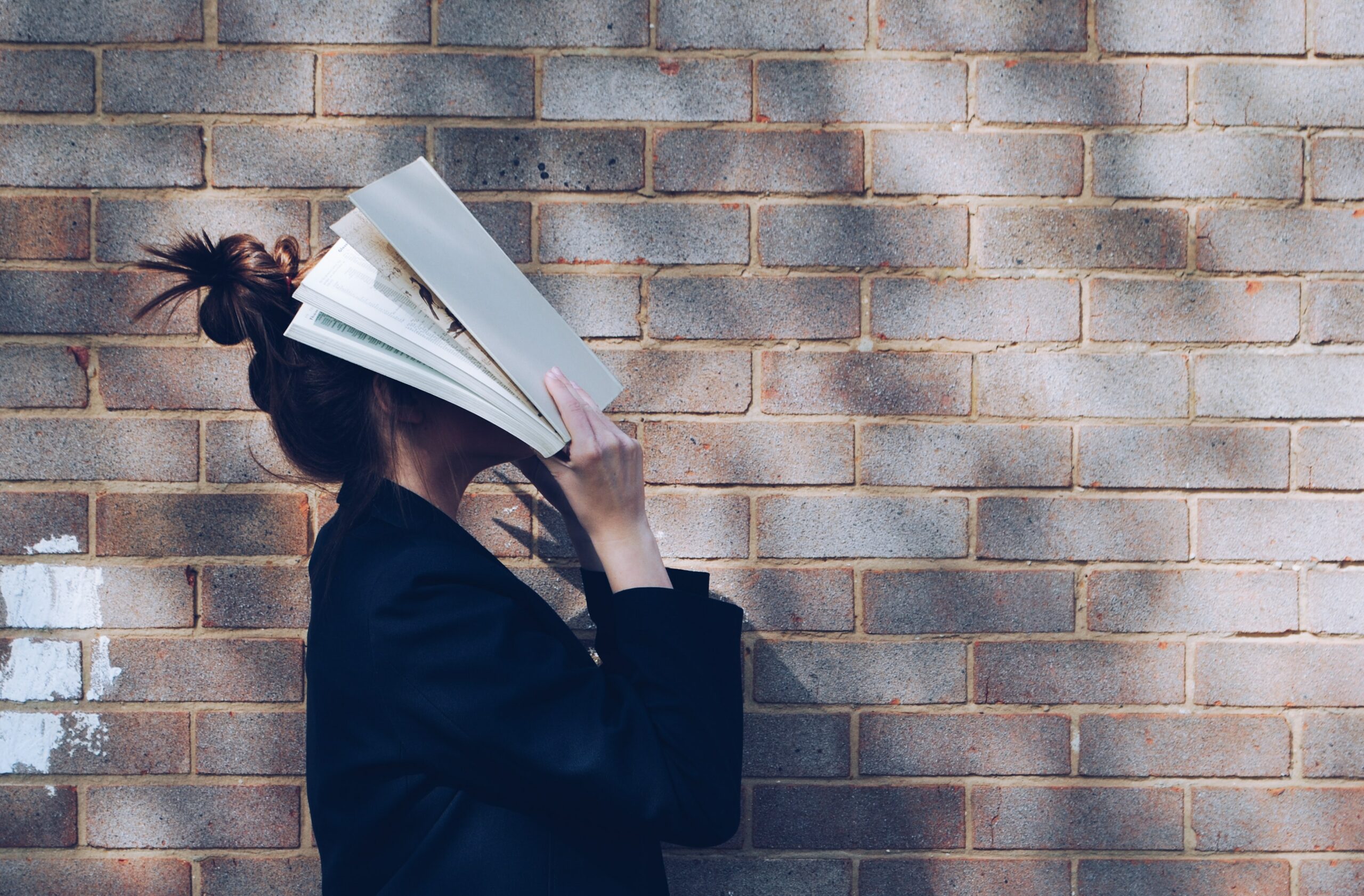 A photo of a woman standing side-on in front of a brick wall. She is wearing a black top, and holding a book right up close to her face, so her face is covered by the book.