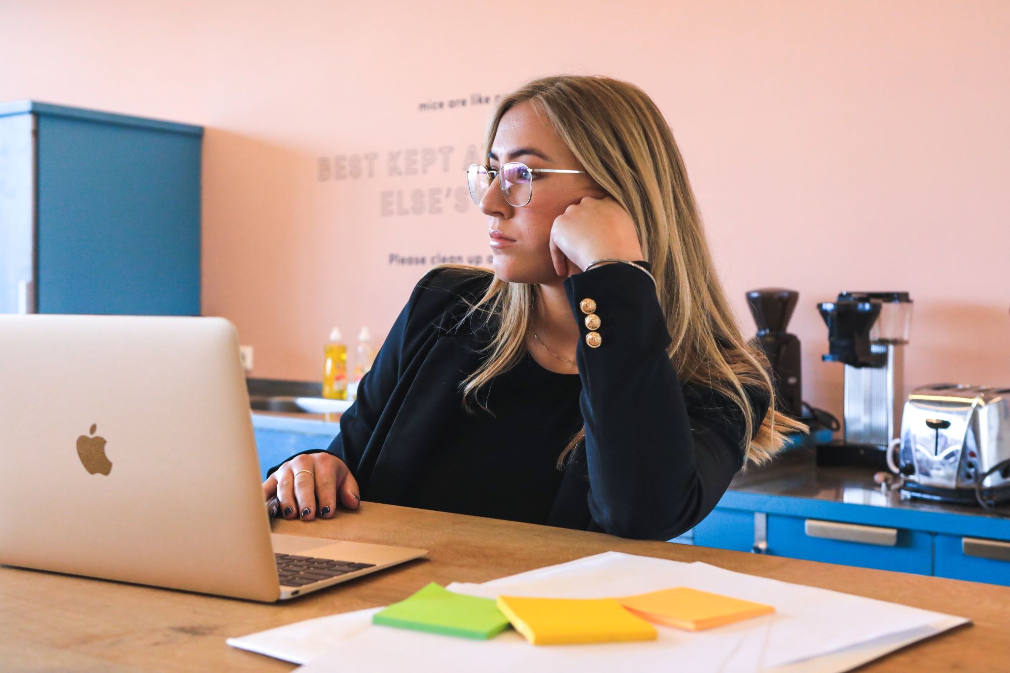 Woman wearing glasses sits at a laptop. She is in a thinking pose. Perhaps she in wondering if what she is experiencing is imposter syndrome, fear or self doubt.