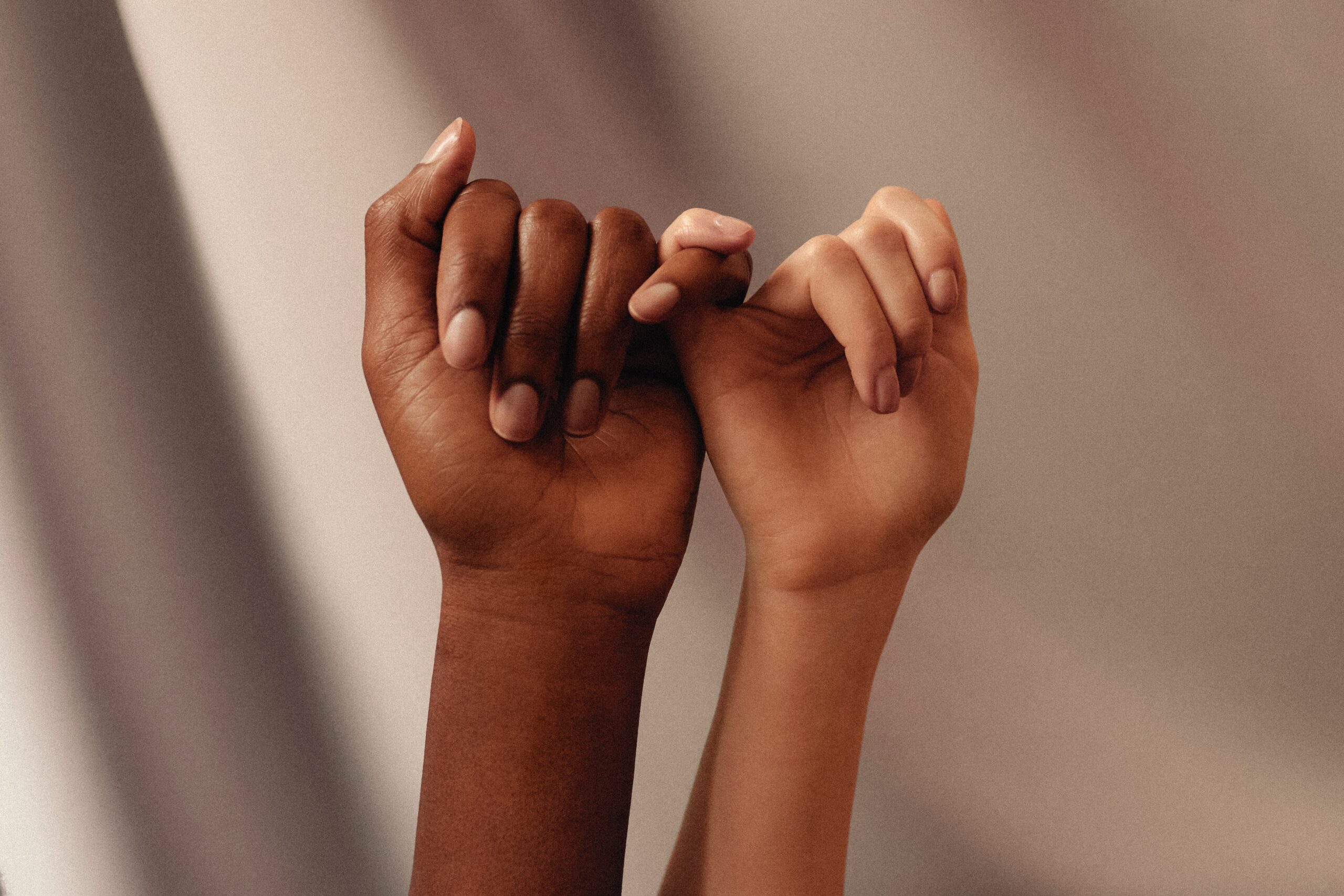 Two women's hands holding pinky fingers. One hand has dark skin and the other hand has lighter skin. Symbolising women working together to break the bias at work.