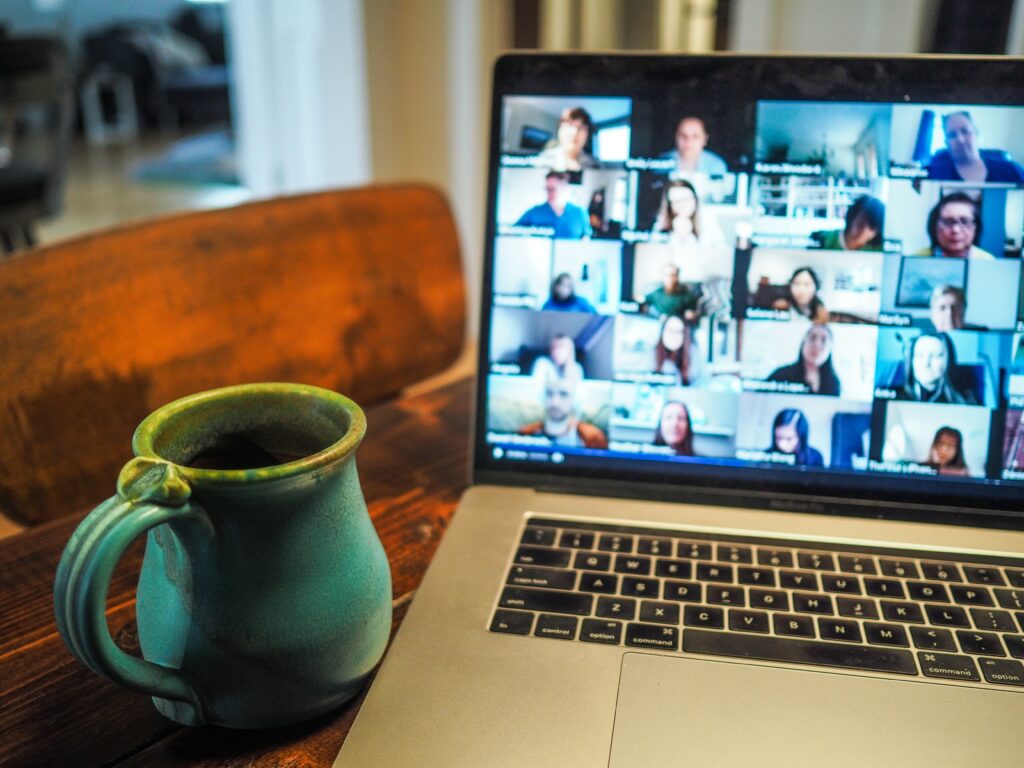Gen Zs are experiencing more social anxiety in the workplace, which can trigger imposter syndrome. [Image description: a laptop showing a Zoom meeting screen sits on a home office desk next to a coffee cup]